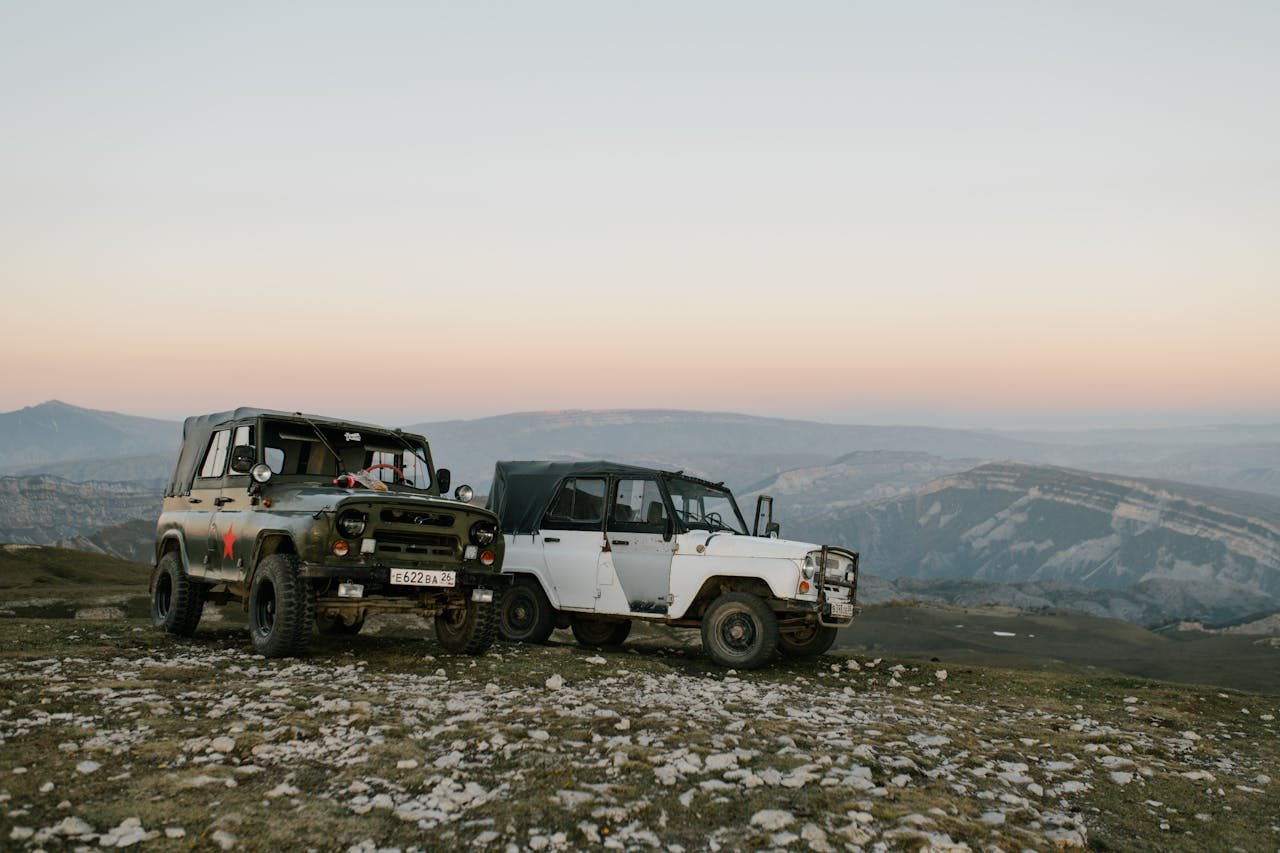 mobile-01 Two vintage off-road vehicles parked on a rocky hill at sunset with a mountain backdrop.